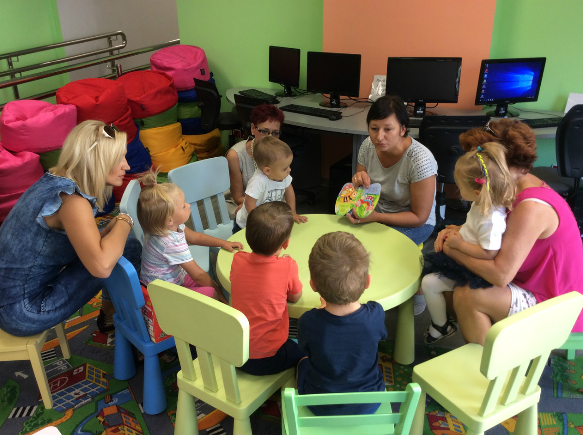 Przy stoliku z bibliotekarką Anią Wiśniewską siedzą dzieci z opiekunkami, słuchają bajki o motylku Kamilku.
