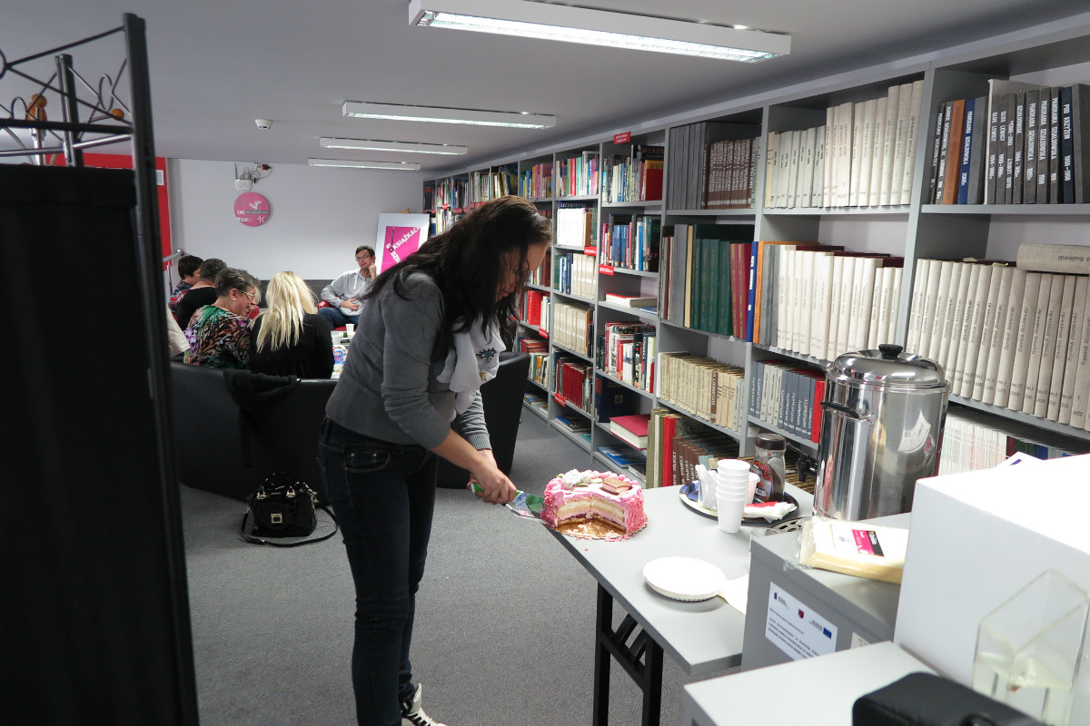 Wnętrze biblioteki, przy stoliku jedna  z uczestniczek DKK CKU kroi tort z okazji pierwszego roku działania klubu.