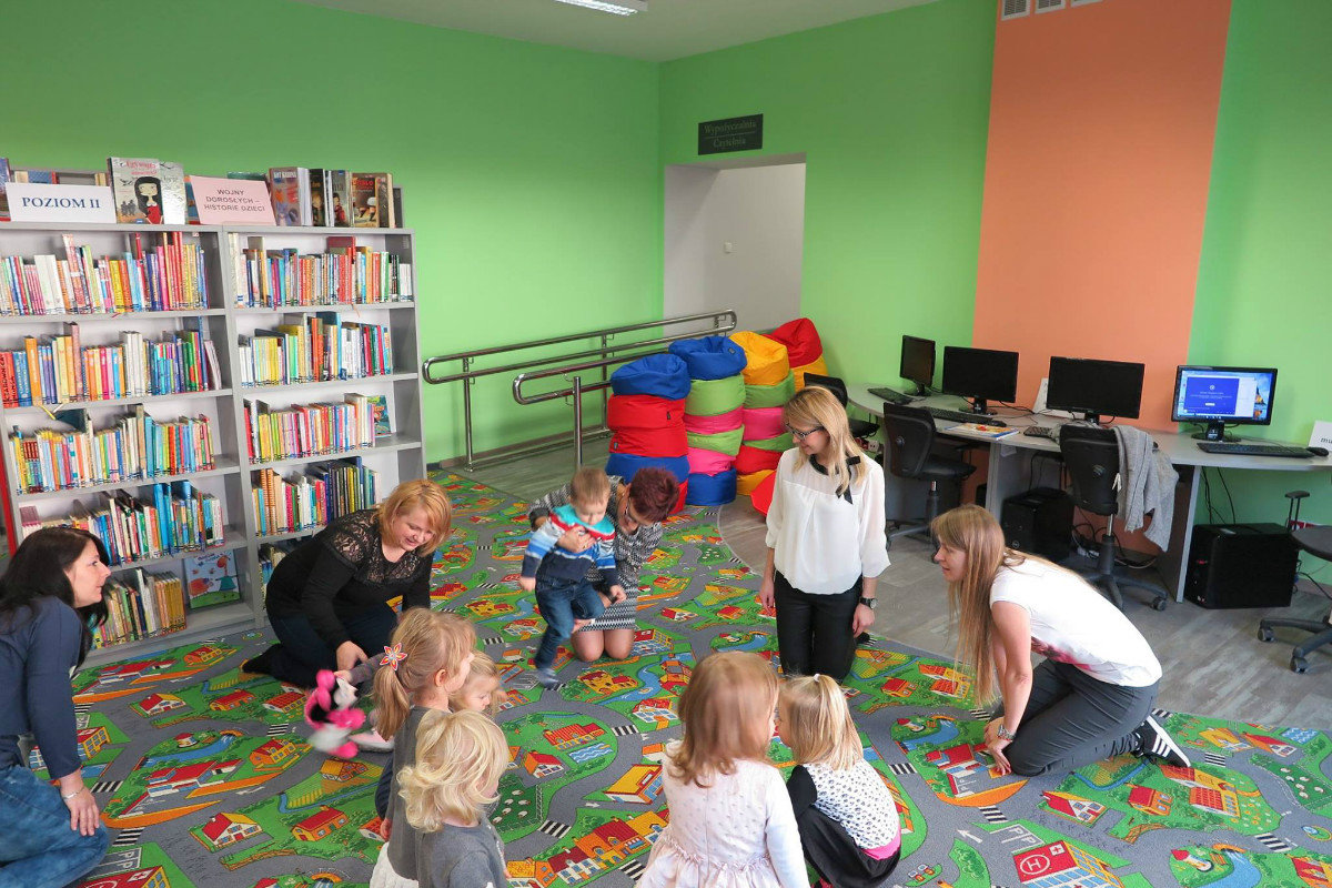 Wnętrze biblioteki. Grupa dzieci z opiekunami podczas zajęć w Klubie Malucha.