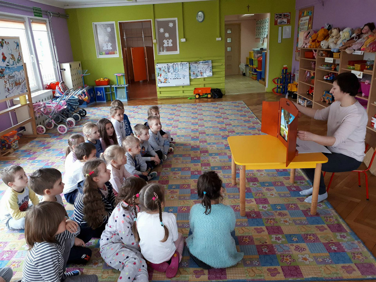 Wnętrze Przedszkola nr 4 na kolorowej wykładzinie siedzą dzieci, bibliotekarka Ania Wiśniewska czyta teatrzyk kamishibai "Legendę o Smoku Wawelskim".