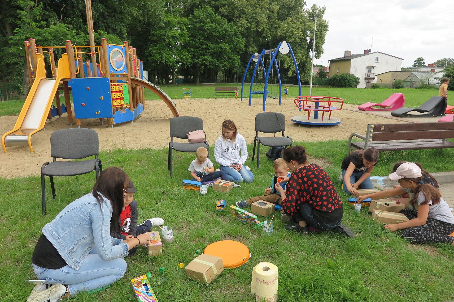 Przy placu zabaw na trawniku siedzi sześcioro dzieci oraz trzech opiekunów malują papierowe ule. 