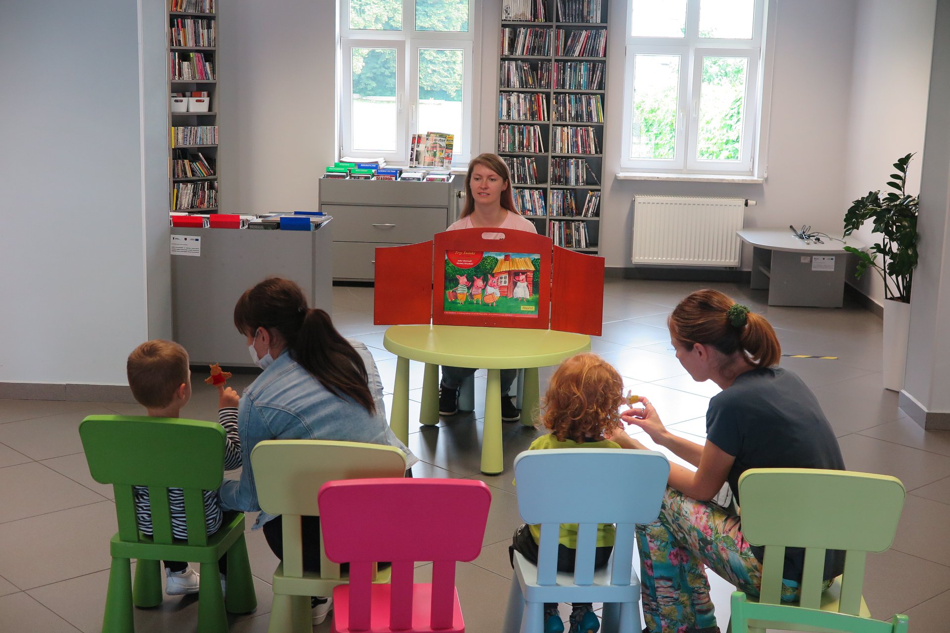 Wnętrze biblioteki mediateka. Bibliotekarka Aleksandra Cybulska przy okrągłym stoliku czyta historię trzech świnek i jednego sprytnego wilka. Dwoje dzieci siedzi na krzesełkach oraz dwóch opiekunów. 