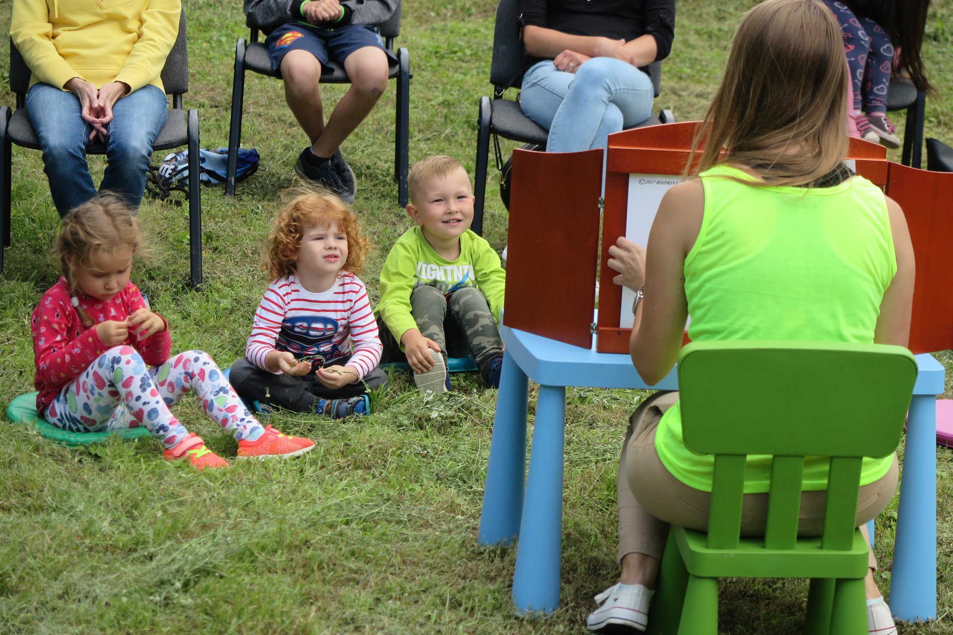 Bibliotekarka Aleksandra Cybulska czyta dzieciom bajkę pt. ,,Przygody pingwina Pingo''. Troje dzieci siedzi na trawie i słucha bajki. Czterech opiekunów siedzi na krzesełkach.