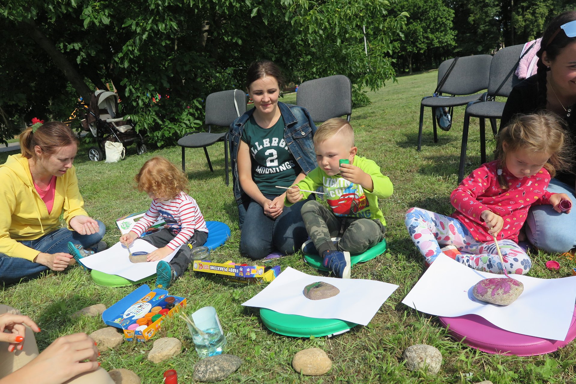 Na trawniku siedzi troje dzieci oraz trzech opiekunów, malują kamienie farbkami.