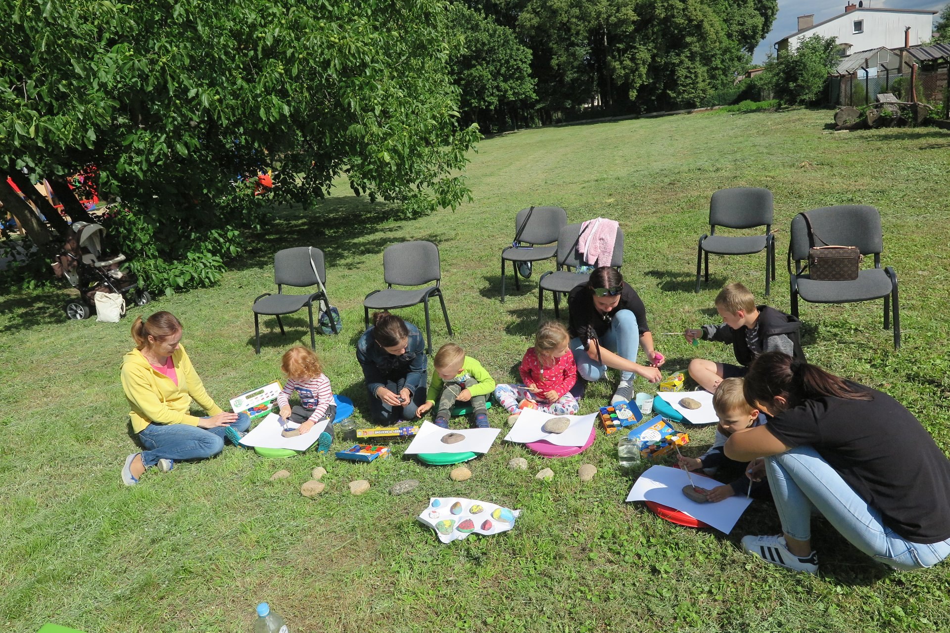 Na trawniku siedzi pięcioro dzieci oraz czterech opiekunów. Dzieci malują kamienie farbkami. 
