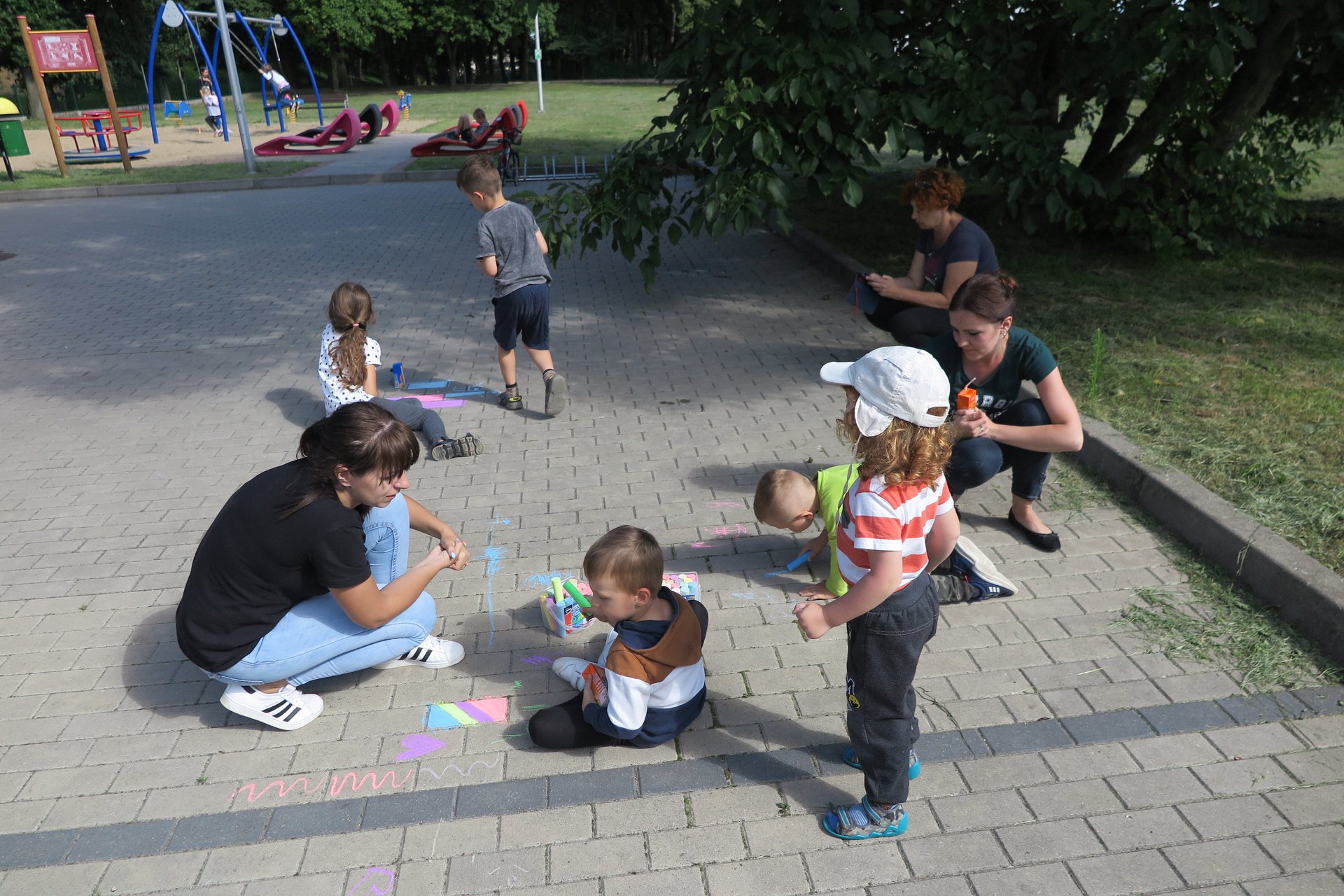 Na placu przed biblioteką pięcioro dzieci bawi się wraz z opiekunami. 