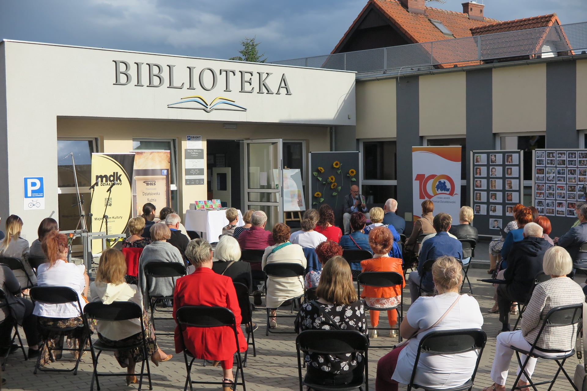 Na placu przed biblioteką w Działdowie z okazji spotkania z poezją zgromadzeni goście siedzą na czarnych krzesełkach. 