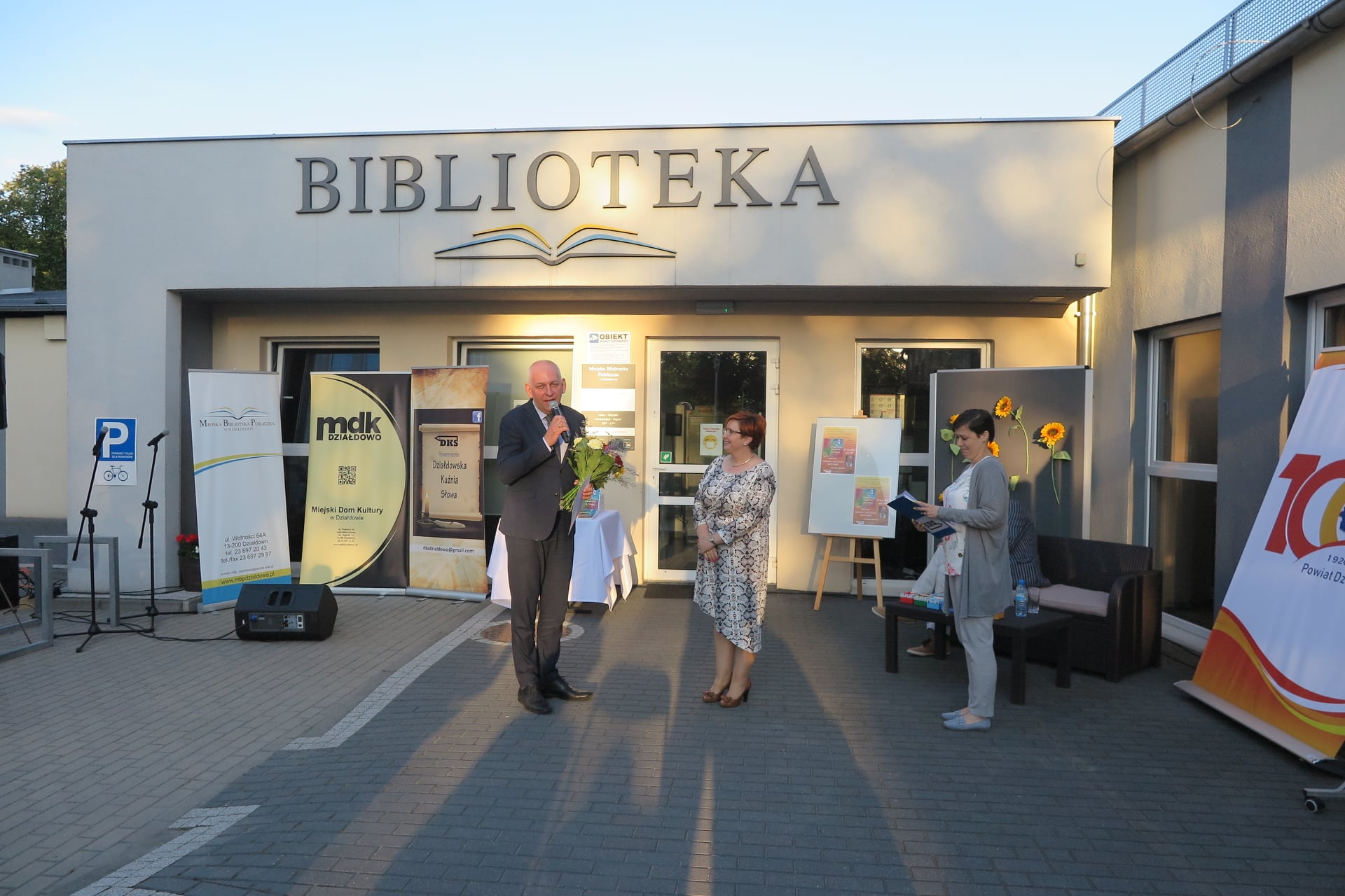 Na placu przed biblioteką z mikrofonem oraz bukietem kwiatów stoi burmistrz miasta Działdowo Grzegorz Mrowiński obok stoją dwie panie z DKS.