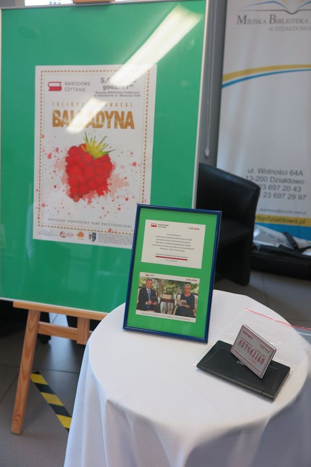 Na stoliku ramka ze zdjęciem Prezydenta Andrzeja Dudy obok sztaluga z plakatem ,,Narodowe Czytanie''. ,,Balladyna''. 