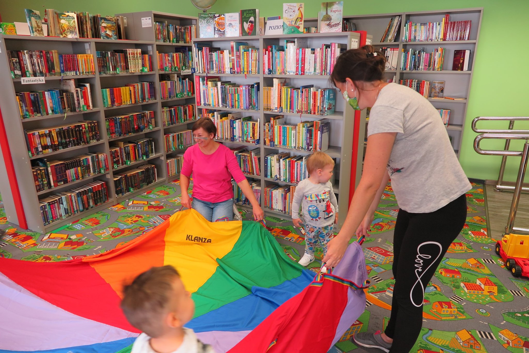 Wnętrze biblioteki (Klub Malucha) zajęcia przeprowadza bibliotekarka Ania Wiśniewska, obok dwoje dzieci wraz z opiekunem. 