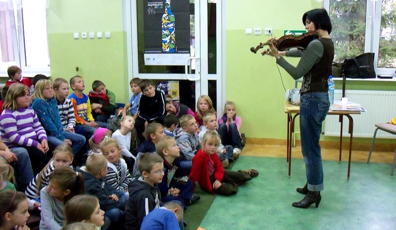 Wnętrze biblioteki. Na skrzypcach gra Izabella Klebańska absolwentka Akademii Muzycznej w Łodzi, w tle regały z książkami i dzieci.
