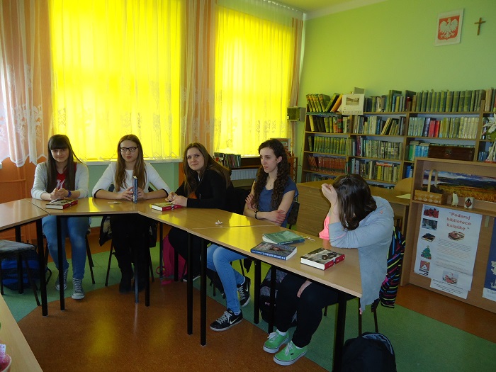 Wnętrze biblioteki, przy stolikach siedzą członkinie DKK GIM NR 2 wspólnie omawiają książkę M. Szczygielskiego "Za  niebieskimi drzwiami". 