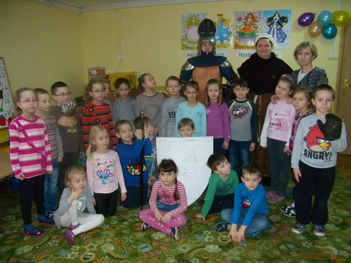 Wnętrze przedszkola, grupa dzieci z opiekunkami i rycerzem z Bractwa Rycerskiego.