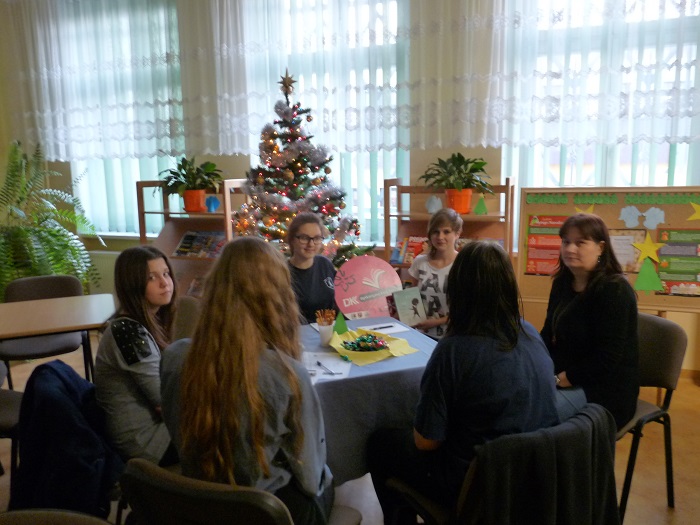 Przy stoliku siedzą uczestnicy DKK GIM NR 1 wspólnie omawiają  książkę F. Moccia "Trzy metry nad niebem" za nimi stoi choinka świąteczna.