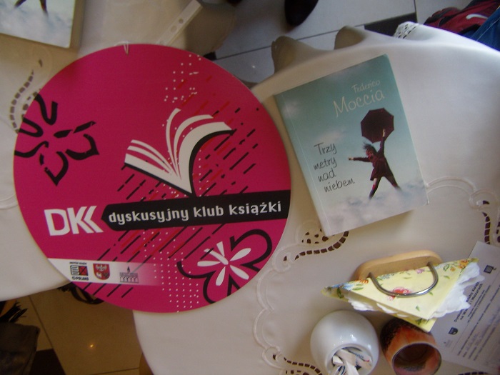 Na stoliku leży okrągłe różowe logo DKK , książka pt. ''Trzy metry nad niebiem'', i serwetnik.
