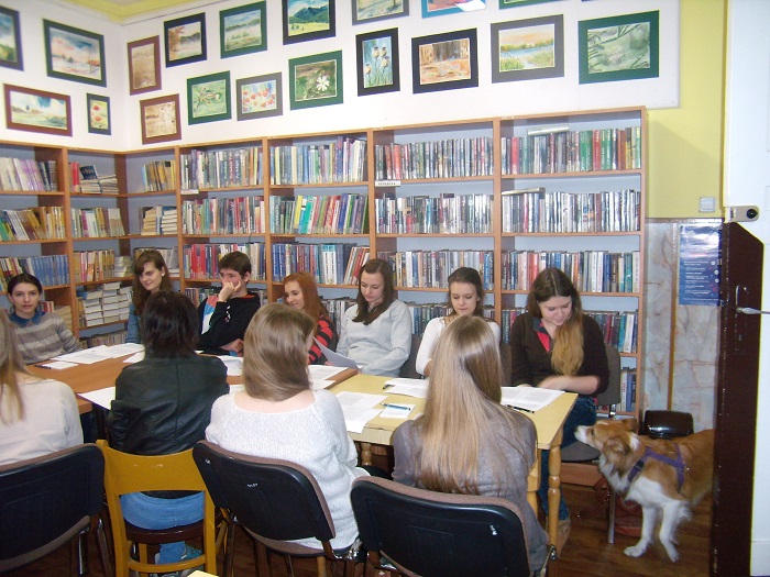 Wnętrze biblioteki, przy stolikach siedzi młodzież za nimi regały z książkami.