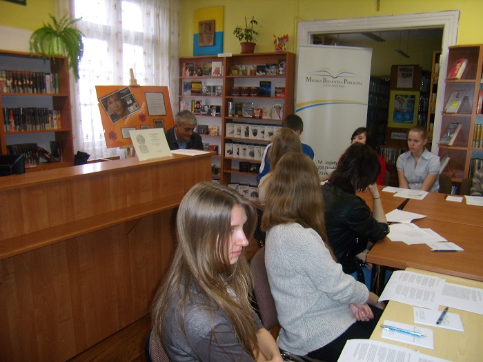 Wnętrze biblioteki, przy stoliku siedzą uczniowie obok Sławomir Matusz prowadzi zajęcia z warsztatów poetyckich.