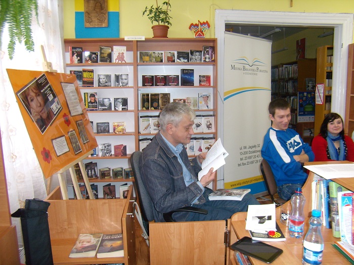 Wnętrze biblioteki, przy stoliku siedzą uczniowie obok Sławomir Matusz prowadzi zajęcia z warsztatów poetyckich.
