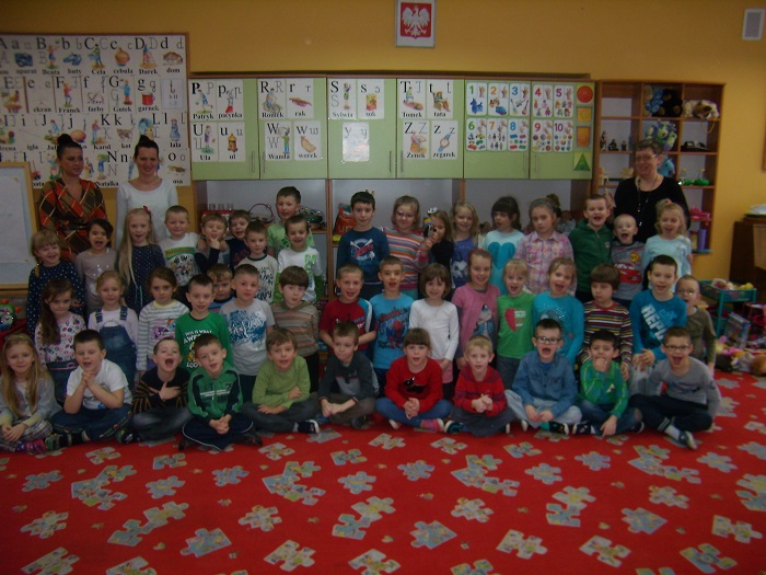 Wnętrze przedszkola, na kolorowej wykładzinie siedzą dzieci , w tle na ścianie wiszą literki poznawane przez dzieci.