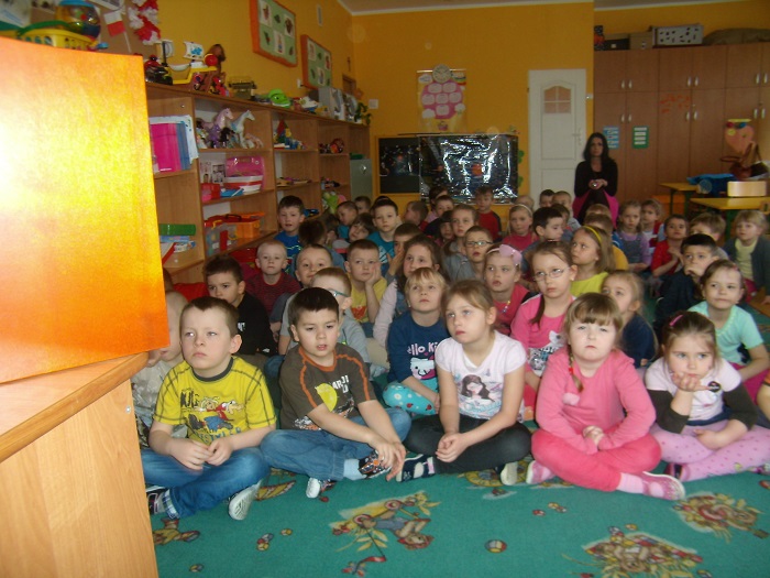 Wnętrze przedszkola, na wykładzinie siedzi grupa dzieci w tle na półkach stoją zabawki.