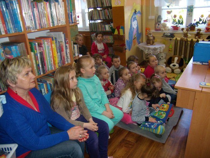 Między regałami z książkami siedzą dzieci biorą udział w lekcji bibliotecznej.