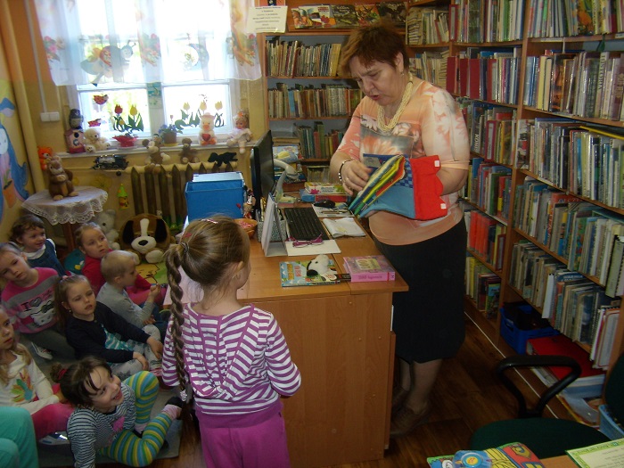 Między regałami z książkami siedzą dzieci biorą udział w lekcji bibliotecznej. 