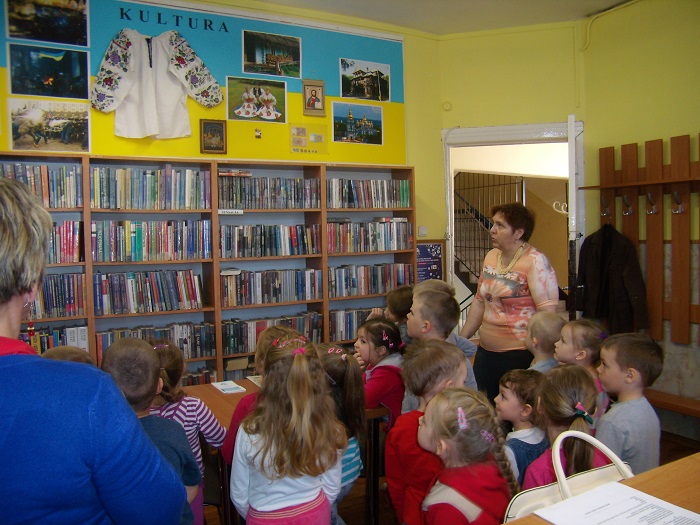 Wnętrze biblioteki, po lewej stronie regały z książkami, obok bibliotekarka i grupa dzieci.