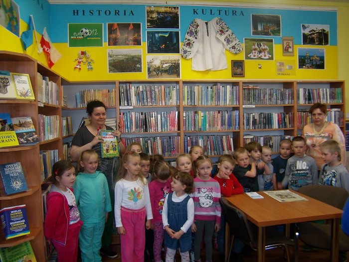 Wnętrze biblioteki, przy ścianie stoją regały z książkami, obok bibliotekarka i grupa dzieci.