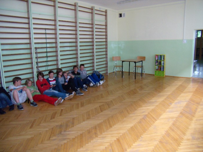 Wnętrze sali gimnastycznej przy drewnianych drabinkach siedzi grupa dzieci.