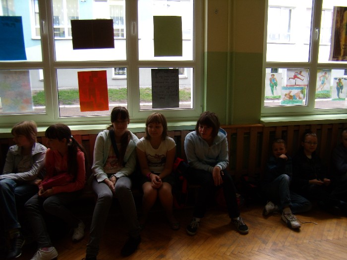 Wnętrze sali gimnastycznej przy oknie na drewnianej ławce siedzi grupa klasowa.
