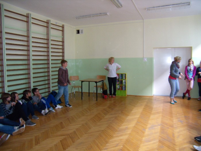 Wnętrze sali gimnastycznej przy drewnianych drabinkach siedzi grupa dzieci w oddali stoją nauczyciele.