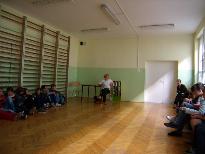 Wnętrze sali gimnastycznej przy drewnianych drabinkach siedzi grupa dzieci w oddali  nauczyciele.
