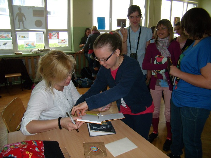 Wnętrze szkoły, przy stoliku siedzi autorka książek dla dzieci i młodzieży Beata Ostrowicka podpisuje młodym czytelnikom swoją książkę.