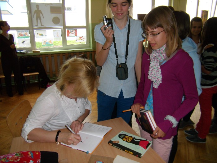 Wnętrze szkoły, przy stoliku siedzi autorka książek dla dzieci i młodzieży Beata Ostrowicka podpisuje młodym czytelnikom swoją książkę.