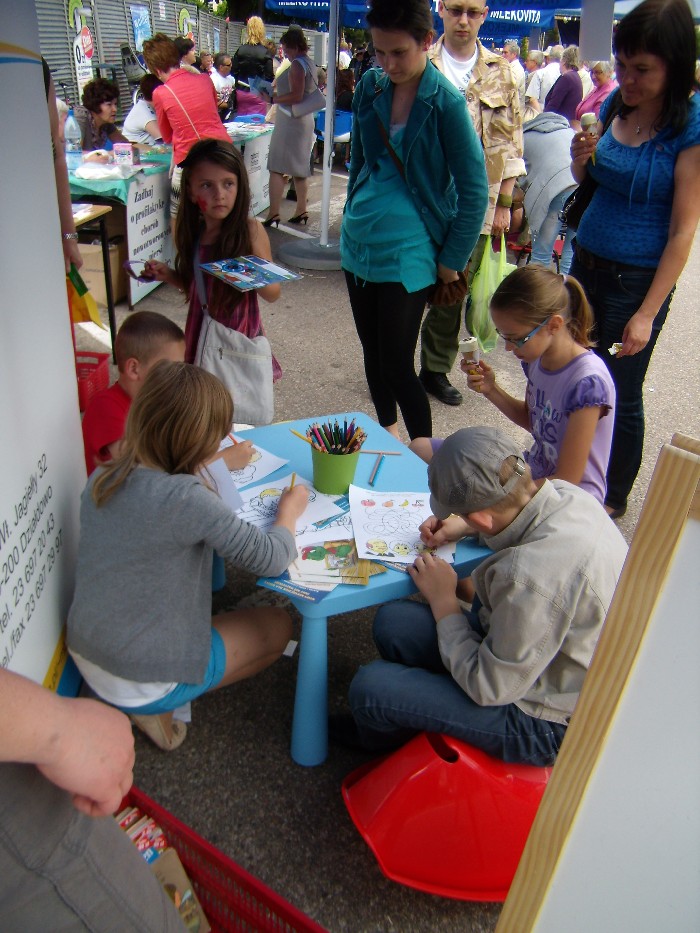 Rynek miasta Działdowa, festyn ''Wstąp po zdrowie'' przy niebieskim stoliku siedzą dzieci wykonują prace plastyczne.