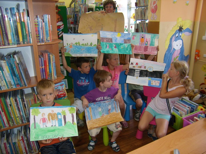 Między regałami z książkami siedzi grupa dzieci wysoko w górze trzyma swoje prace plastyczne na konkurs plastyczny "Bezpieczne Miasto Działdowo".