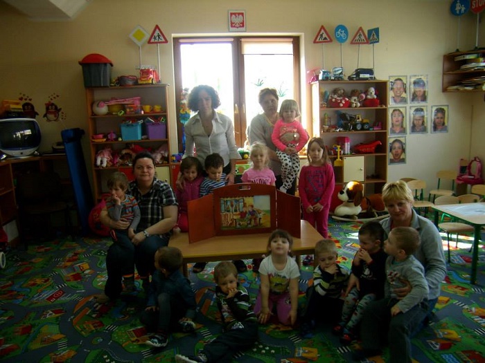 Wnętrze  przedszkola ''Krasnoludek'' przy teatrzyku kamishibai stoi grupa dzieci obok przedszkolanka i bibliotekarka, za nimi regały z zabawkami.
