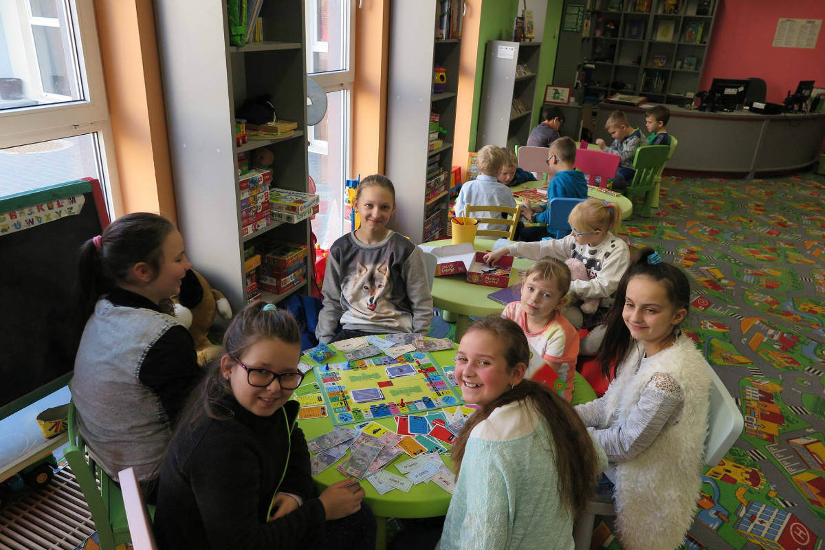 Wnętrze biblioteki. Przy stoliku siedzą dzieci, razem grają w monopol. 
