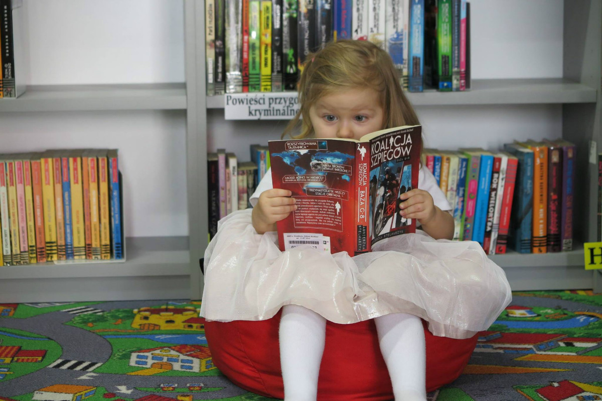 Na czerwonej pufie siedzi dziewczynka z książką pt. ,,Koalicja szpiegów''.