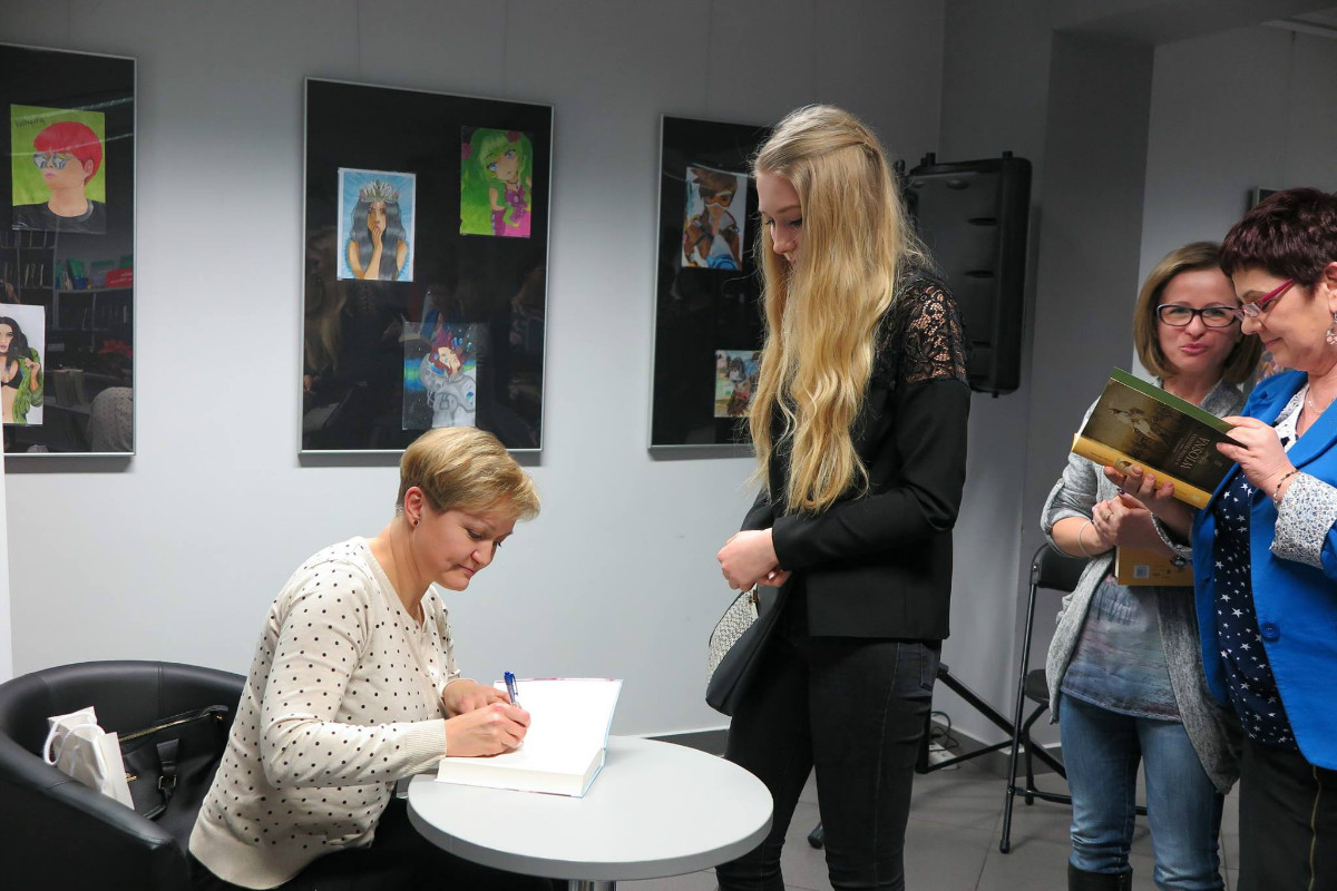 Wnętrze biblioteki,mediateka. Przy stoliku siedzi autorka Agnieszka Olejnik podpisuje książkę młodej czytelniczce.