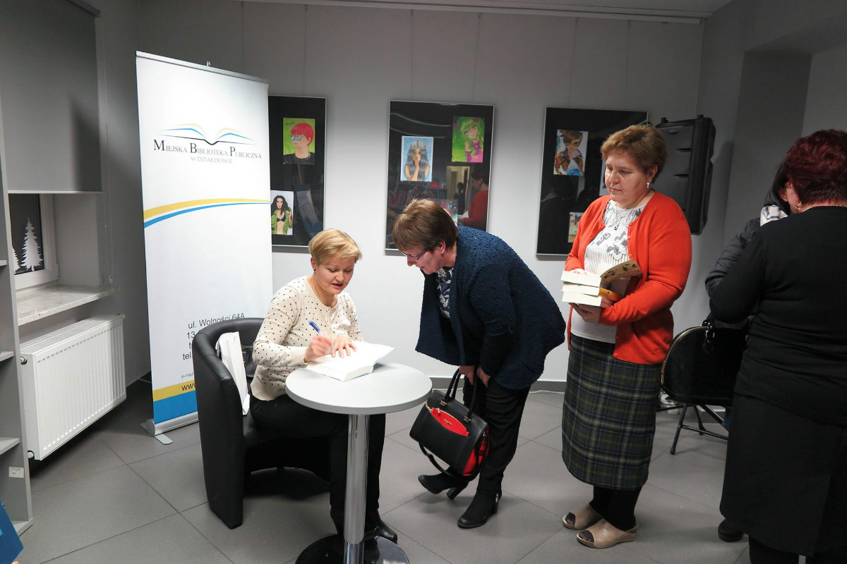 Wnętrze biblioteki,mediateka. Przy stoliku siedzi autorka Agnieszka Olejnik podpisuje książkę  czytelniczce.