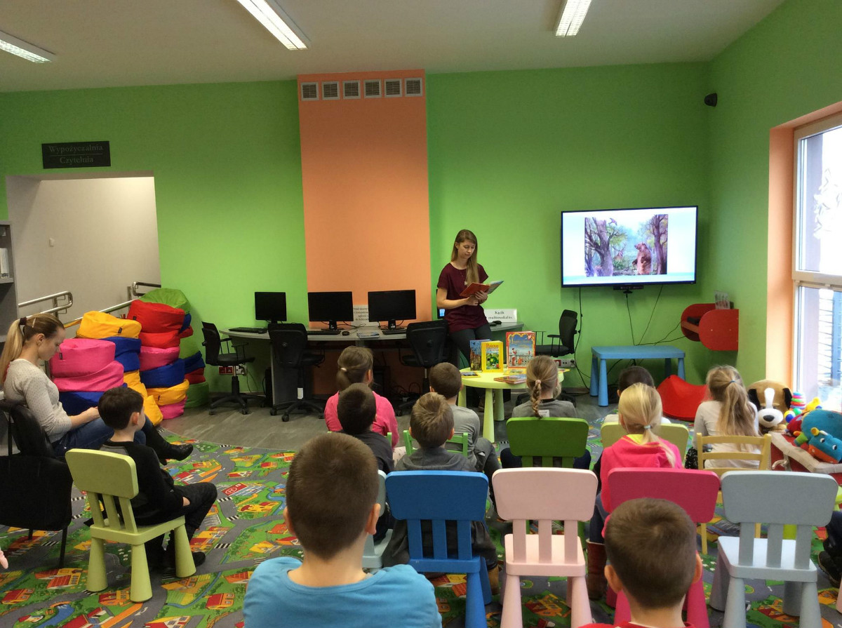 Wnętrze biblioteki, grupa dzieci ze SP nr 4  słucha legendy o toruńskich piernikach, w tle prezentacja na telewizorze.