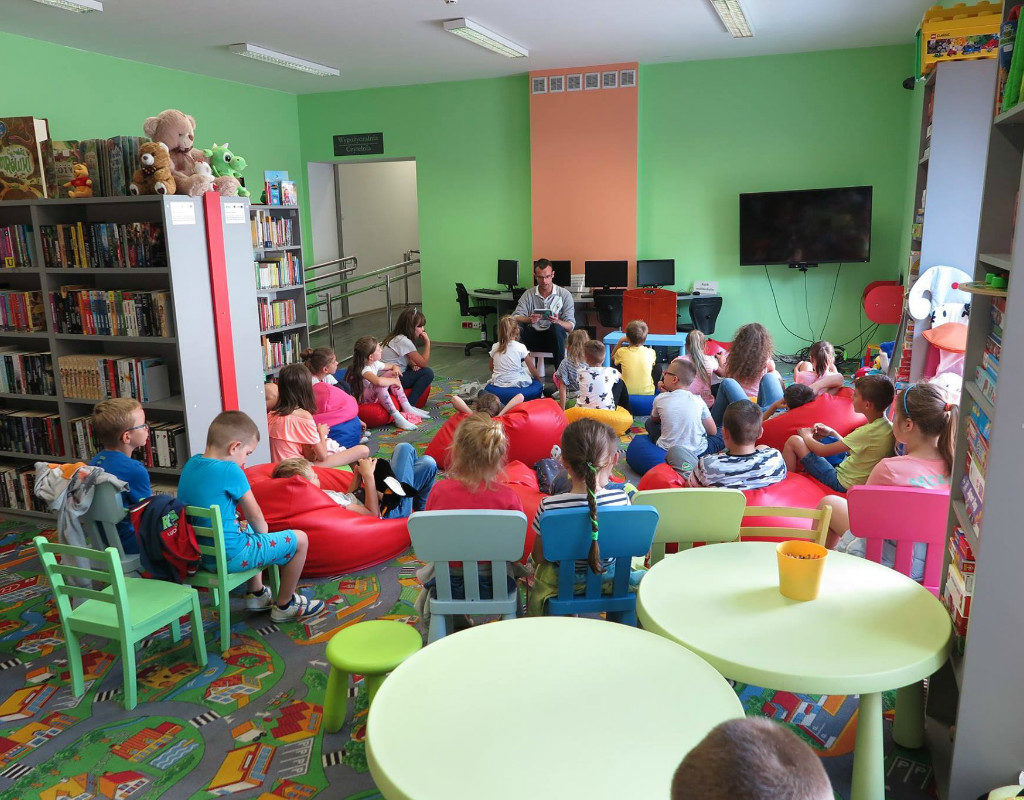 Wnętrze biblioteki, na krzesełkach siedzą dzieci słuchają opowiadania.