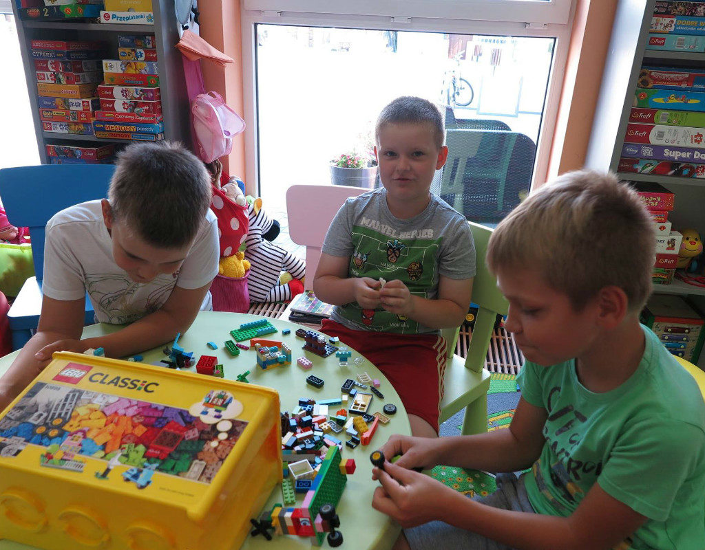 Wnętrze biblioteki. Przy stoliku siedzi trzech chłopców,układają klocki lego.