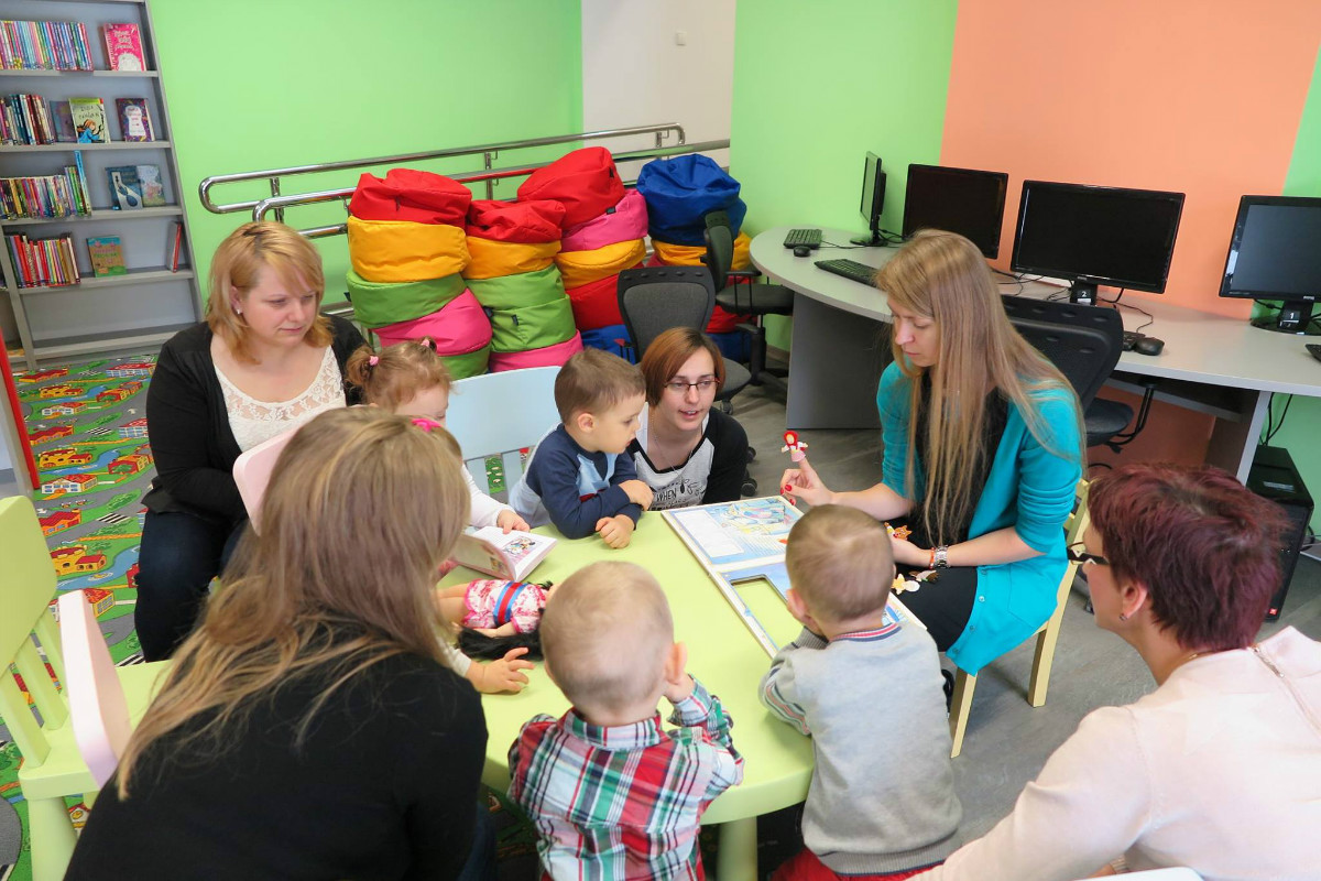 Przy stoliku siedzą dzieci z opiekunami, bibliotekarka Aleksandra Cybulska przedstawia dzieciom teatrzyk paluszkowy o Czerwonym Kapturku.