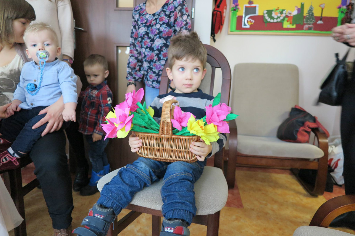 Wnętrze domu seniora. Na krzesełku siedzi chłopiec z koszykiem kwiatów.