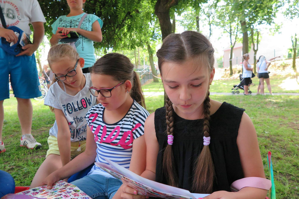 Wnętrze parku, na kolorowej chuście siedzą dziewczynki i czytają książki.