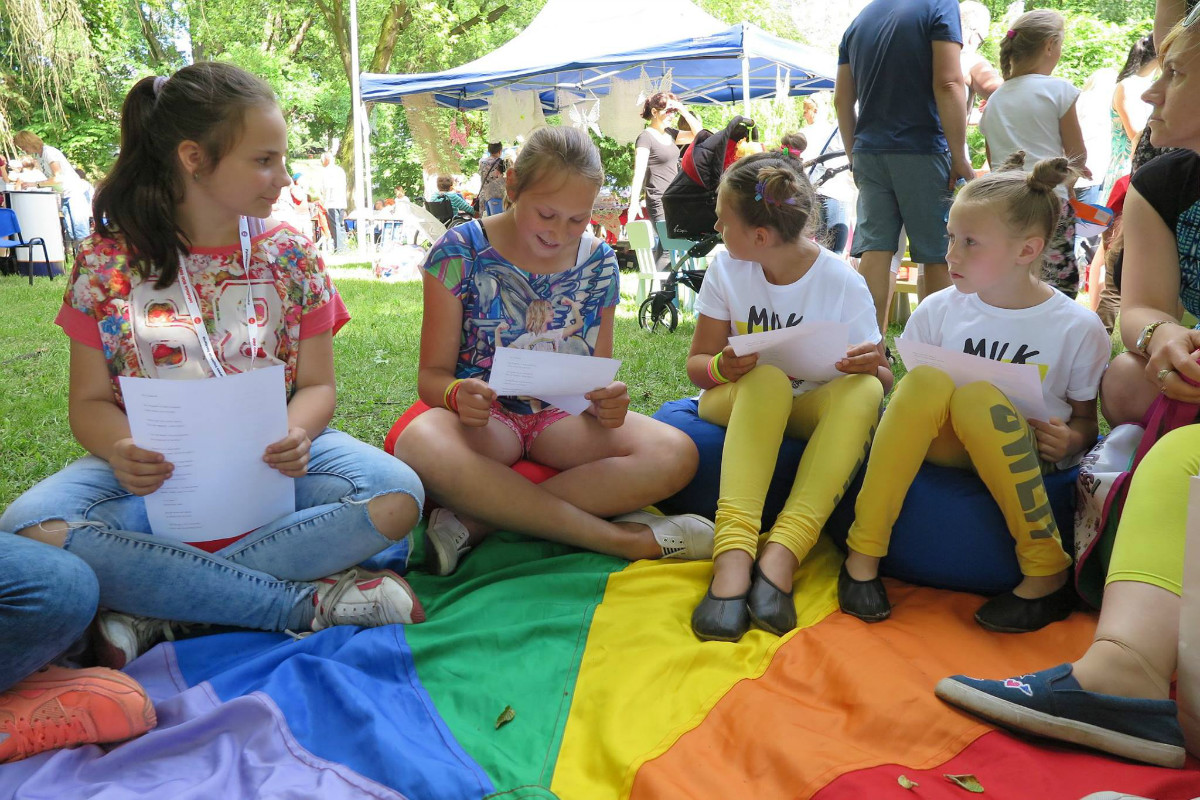 Wnętrze parku, na kolorowej chuście siedzą dzieci i bibliotekarka Ola Cybulska podczas festynu.