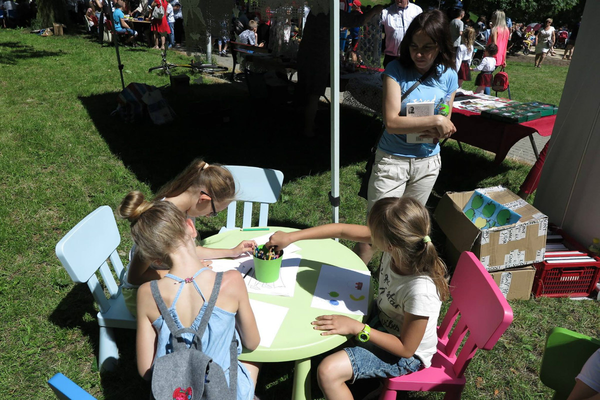 Wnętrze parku. Przy stoliku siedzą trzy dziewczynki razem rysują kolorowymi kredkami. 