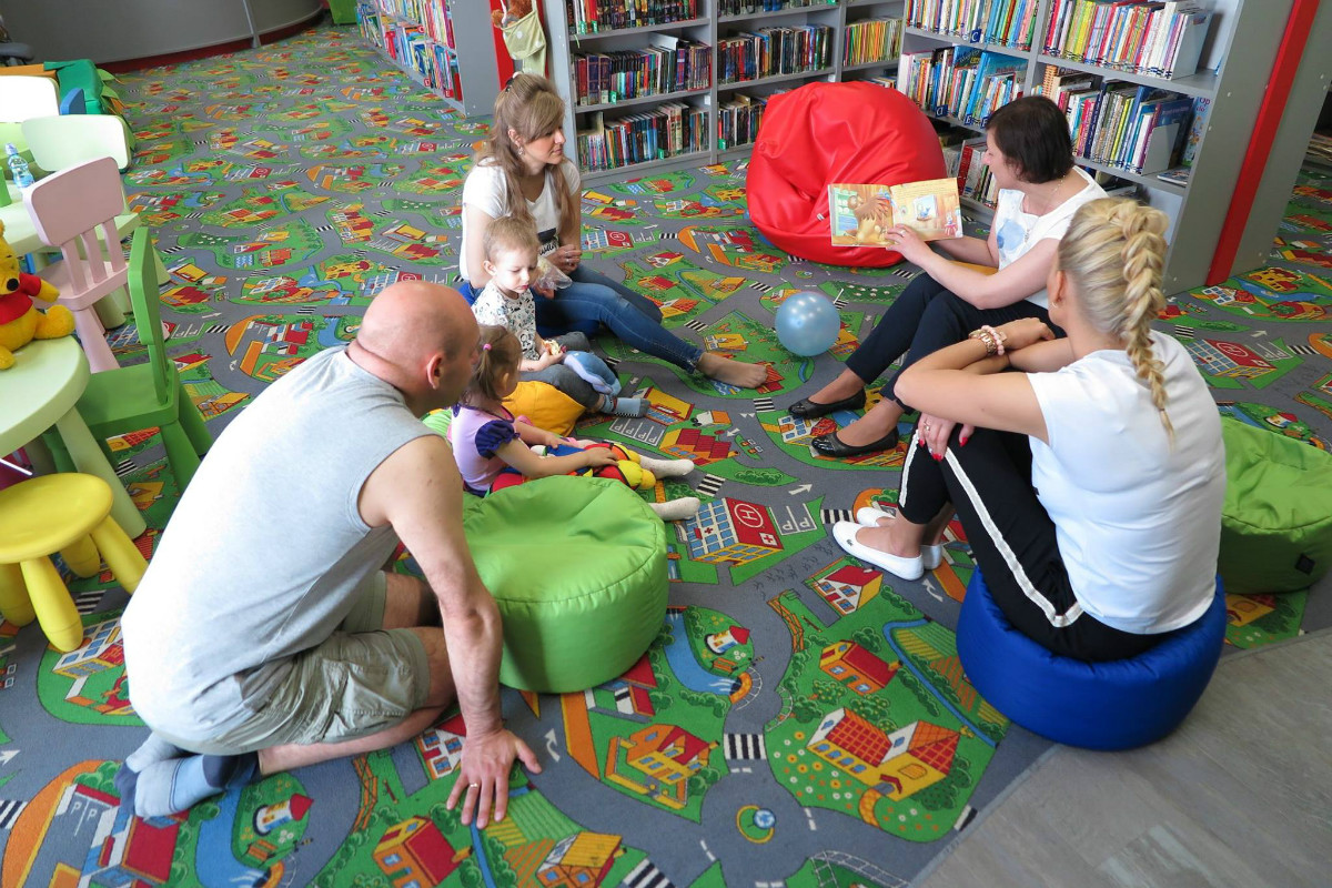 Wnętrze biblioteki. Grupa dzieci z opiekunami słucha opowiadania "Drzewo na prawo, drzewo na lewo" o przygodach Kubusia Puchatka. 